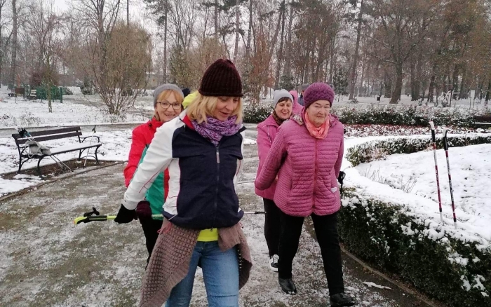 ośnieżony park, aleja parkowa, przy niej ławki grupa osób, na czele 4 kobiety maszeruje trzymając w rękach za sobą kije do nordic walking