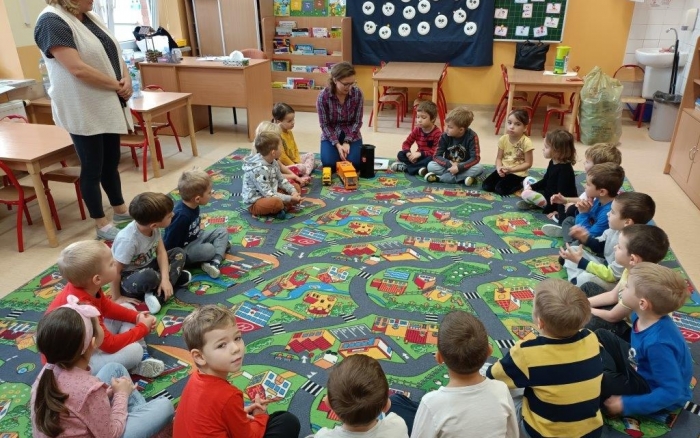sala przedszkolna, grupa dzieci  siedzi na kolorowym dywanie przy nich pracownik urzędu opowiada o prawidłowej segregacji odpadów