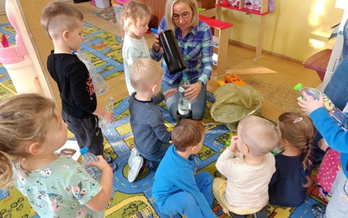sala przedszkolna, grupa dzieci  siedzi na kolorowym dywanie przy nich pracownik urzędu pokazuje zgniatarkę do butelek