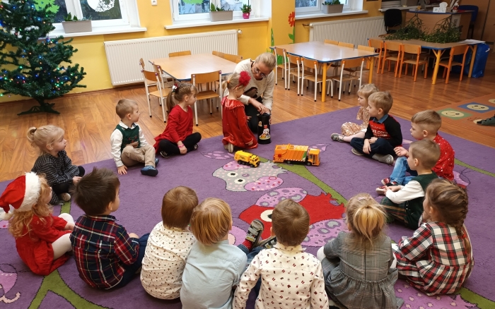 sala przedszkolna, na kolorowym dywanie siedzą dzieci, przed nimi pracownica urzędu pokazuje jak funkcjonuje zgniatarka