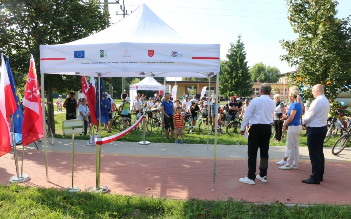 widok na grupę rowerzystów, przed nimi biały namiot przy nim stojaki z 3 flagami: polską, unijną i gminną