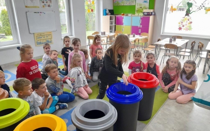 grupa dzieci przedszkolnych w sali dydaktycznej siedzi na kolorowym dywanie, przed nimi ustawione kosze do segregacji odpadów i pracownica urzędu opowiadająca o selektywnej zbiórce odpadów