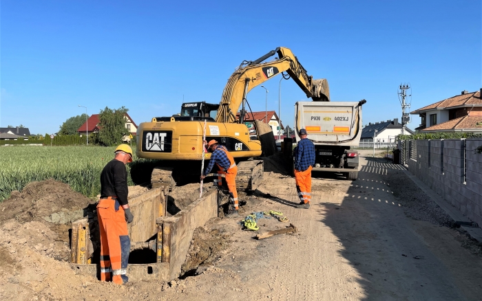 Budowa  kanalizacji sanitarnej w  Gowarzewie  - koparka przesypująca piach na przyczepę, trzech pracowników wykonujących prace budowlane