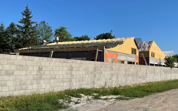 Budowa publicznego przedszkola w Tulcach (inwestor prywatny) - za betonowym ogrodzeniem widok na budowany budynek wykonywane prace przy dachu 